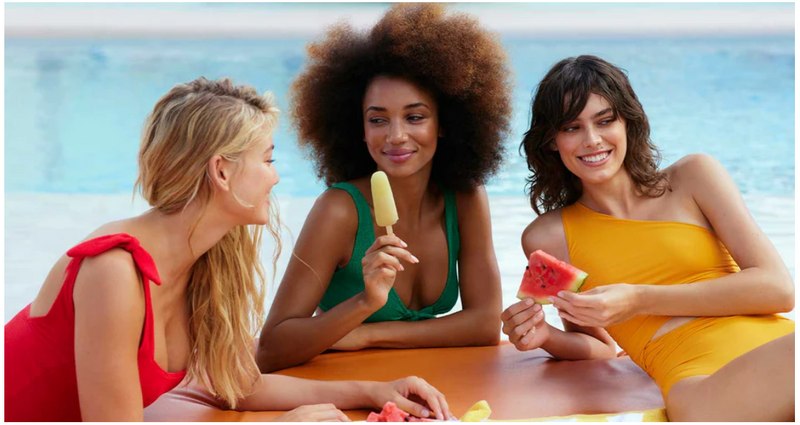 Three women relaxing by the beach in summer, wearing bright swimwear and enjoying fruit, representing effortless summer hair and lifestyle inspiration
