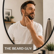A man with a beard and short brown hair looks into a round mirror, touching his chin. He is wearing a white t-shirt in a bathroom setting.