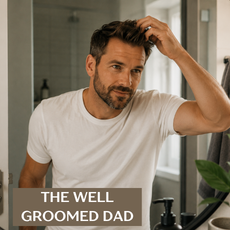 A man with a beard and short hair in a white t-shirt looks into a bathroom mirror, touching his hair. Text overlay at the bottom reads 'THE WELL GROOMED DAD'.