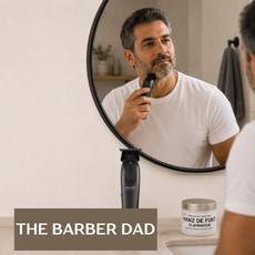 A man in a white t-shirt stands in front of a bathroom mirror holding an electric shaver, with his reflection visible. A jar of beard grooming product sits on the counter below, and a text overlay at the bottom reads 'THE BARBER DAD'.